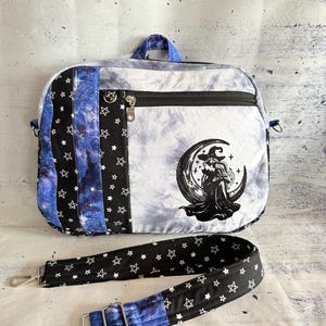 May include: A fabric crossbody bag featuring a tie-dye gray and white main panel with a black embroidered witch sitting on a crescent moon. The side panels display a black fabric with white stars and a blue galaxy print. Includes a detachable strap.