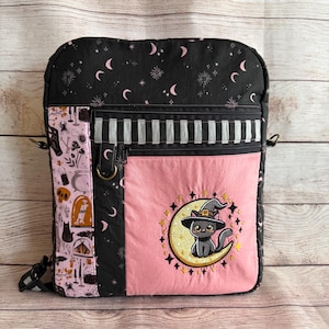 May include: A rectangular backpack featuring a black fabric with pink crescent moons and stars. It includes a side panel with a light pink print of potions and skulls, a black and white striped accent, and a pink front pocket with an embroidered black cat wearing a witch hat sitting on a yellow crescent moon.