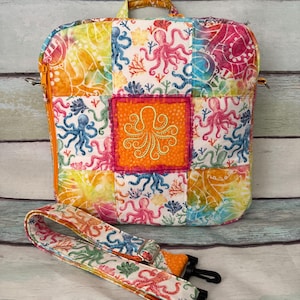 May include: A square quilted bag featuring a colorful octopus and coral print in shades of pink, blue, yellow, and orange. The center panel displays a bright orange square with a stitched octopus design. It includes a top handle and a detachable strap.