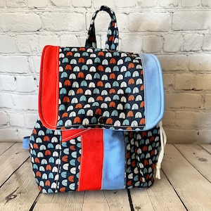 May include: A blue, red, and white patterned backpack with a drawstring closure. The backpack has a flap with a button closure and a single strap.