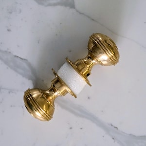 May include: A gold-coloured door knob with a ridged design on each end. The knob is connected by a white foam spacer. The hardware is set against a white and grey marble surface.