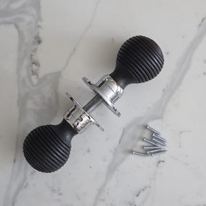 May include: A black and chrome door handle with a ribbed design. The handle has a cylindrical shape with a chrome centre and is accompanied by several small silver screws, suggesting installation hardware. The background is a white marble surface.