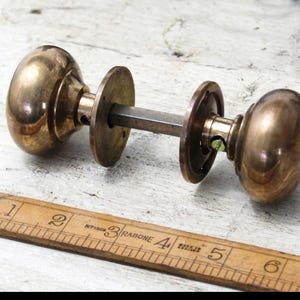May include: Vintage brass door knob set featuring two rounded knobs and circular backplates. The knobs are connected by a square rod. A wooden ruler is placed beside the door knob set.