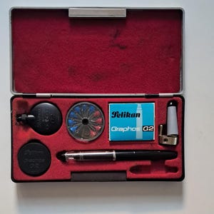 Könnte beinhalten: Geöffnete schwarze Box mit rotem Samtfutter, enthält einen schwarzen Pelikan Graphos G2 Füllfederhalter, Tintenpatronen und Zubehör. Die Box enthält eine kleine blaue Schachtel mit dem Pelikan-Logo.