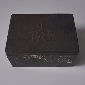 May include: A rectangular, dark brown metal box with a lid. The lid features the text "RIEMSMA SORTE" and the letters "R6". The box has a slightly worn appearance, with some areas showing lighter patches of wear. The overall design is simple and utilitarian.