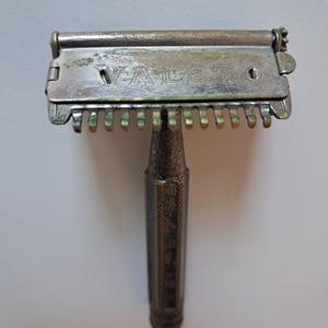 Könnte beinhalten: Vintage silberfarbener Rasierer mit dem Wort "VALET" auf der Oberseite. Der Rasierer hat einen strukturierten Griff und einen klappbaren Kopf mit einem kammartigen Schutz. Das Metall weist Gebrauchsspuren auf.