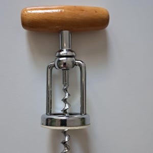 Könnte beinhalten: Ein Korkenzieher aus Chrom und Holz mit einem Holzgriff. Der Metallrahmen hat ein doppeltes Scharnierdesign, und die Schraube hat eine Spiralform. Der Korkenzieher ist zum Entfernen von Korken aus Weinflaschen bestimmt.