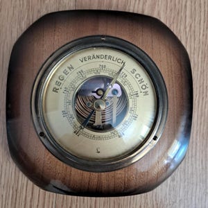 Pode incluir: Um barómetro vintage com moldura de madeira e mostrador cor de latão. O ponteiro indica as condições meteorológicas, com as palavras "REGEN", "VERÄNDERLICH" e "SCHÖN". Mede a pressão atmosférica em milibares.