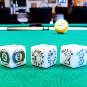 May include: Three white dice with black and blue text and graphics sit on a green pool table. The dice show the numbers 9, 10, 2, and 3, and the phrases "Winner Breaks" and "Loser Shakes". A yellow and white billiard ball is in the background.