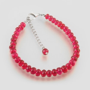 Op de afbeelding: Een armband met kleine, gefacetteerde, rode kralen. Het heeft een zilverkleurige sluiting en een verstelbare ketting. De kralen zijn gelijkmatig verdeeld en vormen een doorlopende lus. De armband is ontworpen om om de pols te worden gedragen.