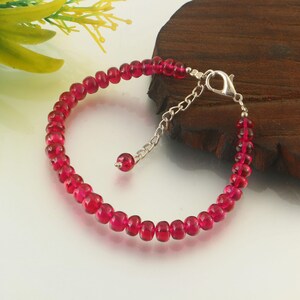 Puede incluir: Una pulsera compuesta por pequeñas cuentas circulares de color rojo intenso y translúcido. La pulsera cuenta con un cierre de mosquetón de metal plateado y una cadena de extensión ajustable, con una cuenta roja a juego al final.