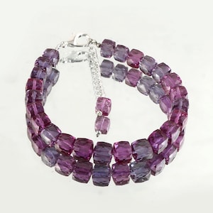May include: A double-strand bracelet featuring faceted, translucent purple and violet cube-shaped beads. The bracelet includes a silver-toned metal chain extender and a lobster claw clasp, with two additional matching beads dangling from the end.
