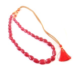 Può includere: Una collana composta da un singolo filo di perline rosso scuro, levigate e di forma irregolare. La collana include un cordino intrecciato color oro con un meccanismo a nodo scorsoio regolabile e una nappa rosso vivo all'estremità.