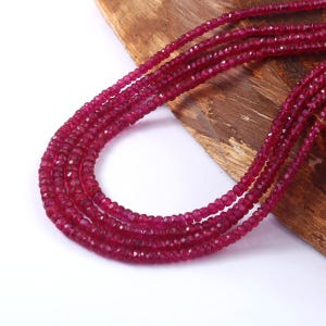 May include: A collection of multiple strands of faceted, deep red ruby beads. The beads are strung together, creating a layered effect. The gemstones are displayed on a wooden surface.