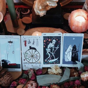 Könnte beinhalten: Vier Tarotkarten, die in einer Holzkiste mit getrockneten Blumen und Tannenzapfen ausgestellt sind. Die Karten zeigen Illustrationen von Figuren und Symbolen. Eine brennende Kerze befindet sich in einem rosa Salzhalter. Die Karten sind beschriftet.