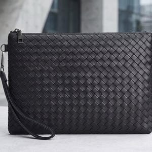 Könnte beinhalten: Schwarze Clutch aus geflochtenem Leder mit Handschlaufe. Die rechteckige Tasche hat einen Reißverschluss und ein Korbgeflechtmuster. Der Riemen ist mit einem silberfarbenen Metallverschluss befestigt. Der Hintergrund ist unscharf.