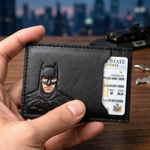 Op de afbeelding: Zwart leren portemonnee met een reliëf Batman-ontwerp. De portemonnee bevat een staatslicentie met de tekst "STATE LICENSE" en andere details. De portemonnee wordt in de hand van een persoon gehouden.