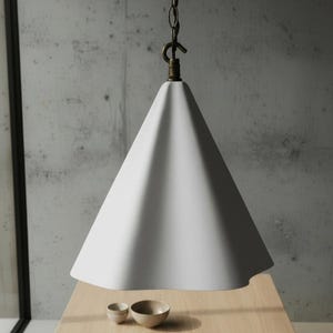 Op de afbeelding: Een witte, kegelvormige hanglamp die aan een ketting is opgehangen. Het licht hangt boven een houten tafel met twee kleine kommen. De achtergrond is een betonnen muur.