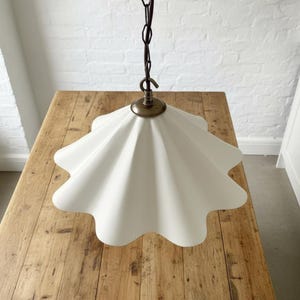 May include: A white, scalloped-edge pendant light fixture. The light has a brass-colored top and a dark brown chain. The fixture is hanging above a wooden table. The background is a white brick wall.