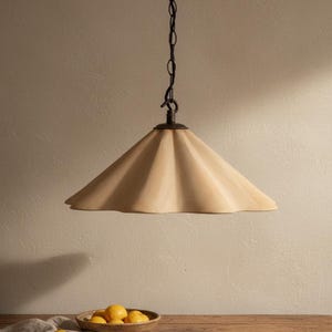 Op de afbeelding: Een beige, kegelvormige hanglamp met een geschulpte rand. Het licht hangt aan een donkere ketting en een cirkelvormige fitting. Het licht hangt boven een houten tafel met een kom met citroenen.