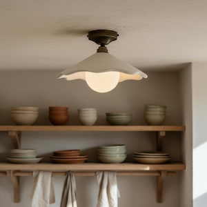 May include: A ceiling light fixture with a white, scalloped shade and a round, glowing bulb. The fixture is mounted on a brass-colored base. Below, wooden shelves display various ceramic bowls and plates in neutral tones, creating a warm, inviting kitchen scene.