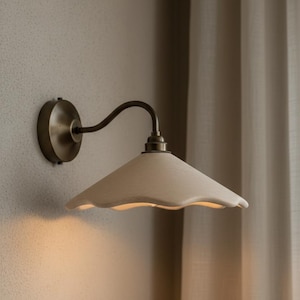 Könnte beinhalten: Wandleuchte mit cremefarbenem, gewelltem Schirm. Die Leuchte hat einen gebogenen, dunklen Metallarm und eine kreisförmige Basis. Die Metallkomponenten haben eine Messingoberfläche. Das Licht ist an einer neutralfarbenen Wand montiert.