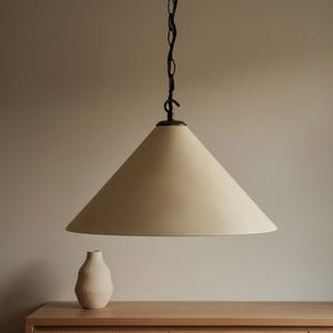 Può includere: Lampada a sospensione a forma di cono color crema sospesa a una catena nera. La luce è sospesa sopra una superficie in legno, con un piccolo vaso strutturato sulla superficie. La lampada ha un design minimalista.