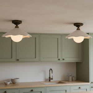 May include: Two white, scalloped-edge pendant lights with brass fixtures hang from the ceiling. Each light has a round, glowing bulb. The lights are above a kitchen with sage green cabinets and a wooden countertop.