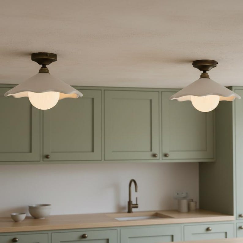 May include: Two white, scalloped-edge pendant lights with brass fixtures hang from the ceiling. Each light has a round, glowing bulb. The lights are above a kitchen with sage green cabinets and a wooden countertop.