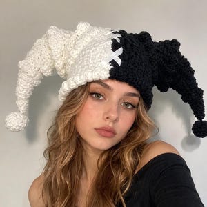 May include: A two-toned jester-style hat crafted from crochet yarn, featuring a split design with one side in solid white and the other in solid black. The hat includes two long, pointed, drooping sections ending in pom-poms and a white ribbon lace-up.