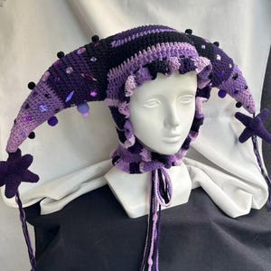 May include: A crochet jester hat in shades of dark purple, light purple, and black. The hat features two long, pointed side panels adorned with small black pom-poms and various purple and clear faceted beads. A star-shaped charm hangs from each tip.