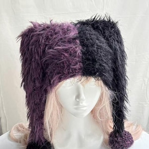 May include: A two-toned fuzzy hat featuring a split design with one side in deep purple and the other in black. The hat has long, dangling earflaps on both sides, each finished with a round, textured pom-pom at the bottom of the long panels.