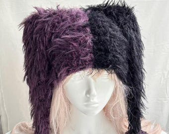 Könnte beinhalten: Eine zweifarbige, flauschige Mütze mit geteiltem Design, wobei eine Seite in Dunkelviolett und die andere in Schwarz gehalten ist. Die Mütze hat lange, herabhängende Ohrenklappen, die jeweils mit einem runden, strukturierten Bommel enden.