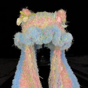 May include: A fuzzy, textured hat in pastel blue and pink with long, dangling side panels. The hat features cat-like ears adorned with small white tulip-shaped ornaments, pink ribbons, and thin, trailing decorative yarn strands throughout.
