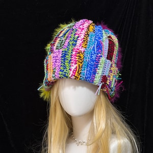 May include: A multi-coloured crochet beanie featuring a vibrant patchwork of vertical stripes in shades of blue, pink, yellow, green, and purple. The hat is accented with fuzzy, textured yarn trim in bright neon yellow and magenta along the edges.