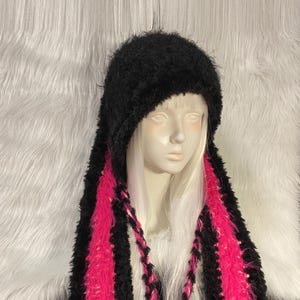 May include: A black fuzzy hat with long ear flaps featuring bright pink centres. The hat includes two braided ties made of black and pink yarn that hang down from the sides. The texture of the material is shaggy and soft, covering the head.