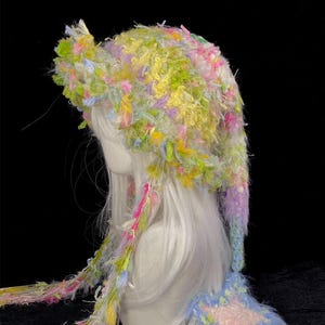 May include: A multi-colored, fuzzy, textured hat featuring a long, pointed tail that ends in a star-shaped ornament. The hat is crafted from yarn in shades of yellow, green, pink, and blue, with long, dangling fringe strands extending from the sides.
