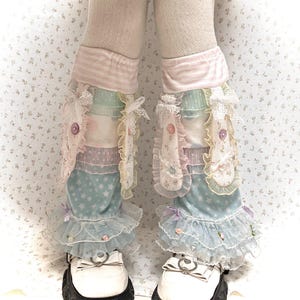 Pode incluir: Um par de polainas em camadas com tecidos em tons pastel rosa, azul e branco com padrões de estrelas e flores. As polainas são adornadas com renda franzida, pequenos laços de tecido, botões decorativos e minúsculas rosas aplicadas.