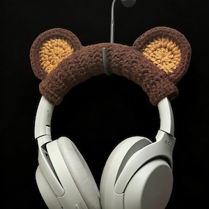 Può includere: Cuffie wireless bianche con fascia per la testa a orecchie di orso all'uncinetto marrone. La fascia ha orecchie interne color cuoio. Le cuffie sono appese a un gancio grigio su sfondo nero. Le cuffie hanno la scritta "SONY".