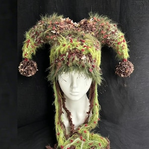 May include: A handmade jester hat with a distinctive, textured design. The hat combines green, brown, and red yarn, with fluffy accents and dangling strands. Two brown pom-poms are attached to the ends of the hat's horns.