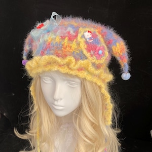 May include: A fuzzy, multi-colored knit hat featuring a yellow ruffled brim, two long earflaps with star-shaped ends, a light blue ribbon bow, and a small Hello Kitty character charm attached to the side. The hat has a soft, textured appearance.