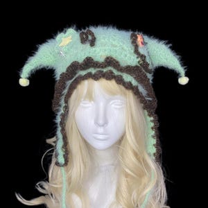 May include: A fuzzy mint green jester-style hat with two pointed sides, each tipped with a small cream pom-pom. The hat features a dark brown scalloped trim along the edge and two long braided ties ending in mint green star-shaped accents at the bottom.