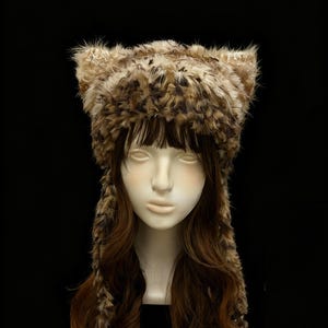 May include: A faux fur hat with pointed ears, in shades of brown and tan. The hat is displayed on a mannequin head with long, wavy brown hair, against a black backdrop. The hat's texture appears soft and fluffy, resembling animal fur.