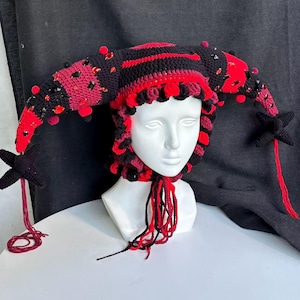 Pode incluir: Um gorro de bobo da corte em croch&ecirc; preto e vermelho com dois chifres longos e curvos adornados com pequenos pompons vermelhos e cristais facetados pretos e vermelhos. O gorro tem uma borda franzida e longas borlas de l&atilde; penduradas.