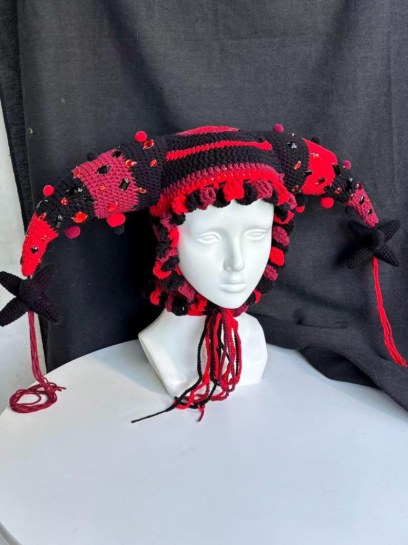 Pode incluir: Um gorro de bobo da corte em croch&ecirc; preto e vermelho com dois chifres longos e curvos adornados com pequenos pompons vermelhos e cristais facetados pretos e vermelhos. O gorro tem uma borda franzida e longas borlas de l&atilde; penduradas.