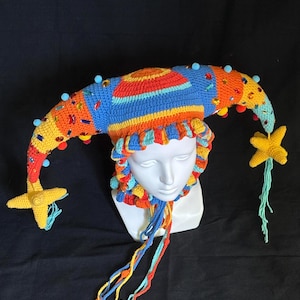 Pode incluir: Um chap&eacute;u de bobo da corte em croch&ecirc; multicolorido com duas pontas longas e curvas adornadas com pequenas esferas azuis, strass coloridos e detalhes em forma de estrela amarela nas extremidades. O chap&eacute;u inclui uma borda franzida e la&ccedil;os.