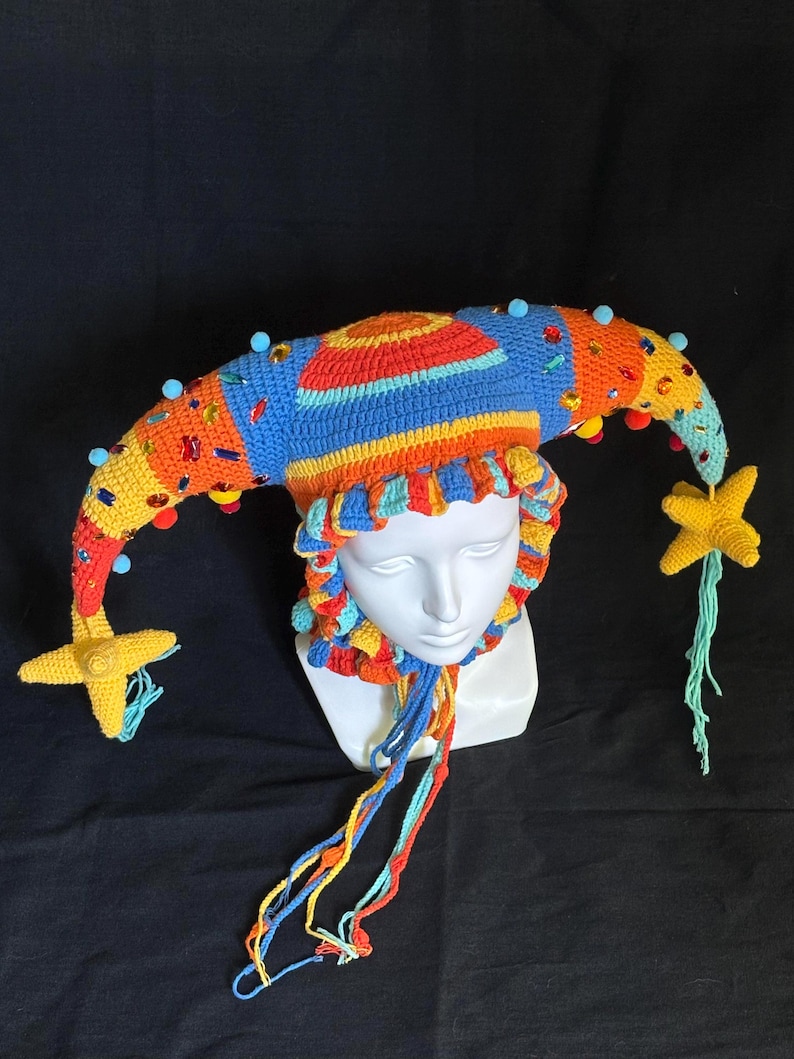 Pode incluir: Um chap&eacute;u de bobo da corte em croch&ecirc; multicolorido com duas pontas longas e curvas adornadas com pequenas esferas azuis, strass coloridos e detalhes em forma de estrela amarela nas extremidades. O chap&eacute;u inclui uma borda franzida e la&ccedil;os.