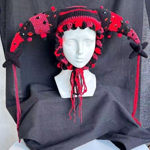 Pode incluir: Um gorro de bobo da corte em croch&ecirc; preto e vermelho com dois chifres longos e curvos adornados com gemas facetadas vermelhas e pretas e pequenos pompons. O gorro tem uma borda franzida ao redor do rosto, borlas pendentes e pontas em estrela.
