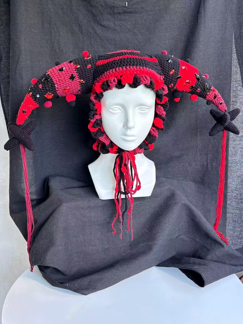 Pode incluir: Um gorro de bobo da corte em croch&ecirc; preto e vermelho com dois chifres longos e curvos adornados com gemas facetadas vermelhas e pretas e pequenos pompons. O gorro tem uma borda franzida ao redor do rosto, borlas pendentes e pontas em estrela.
