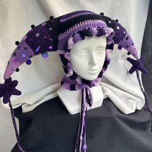 Pode incluir: Um gorro de bobo da corte em croch&ecirc; nos tons roxo escuro, roxo claro e preto. O gorro apresenta dois pain&eacute;is laterais longos e pontiagudos adornados com pequenos pompons pretos e v&aacute;rias contas facetadas roxas e transparentes. Uma estrela pendura.
